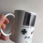Weiße Tasse im Design eines klassischen Nintendo Game Boy mit Bildschirm, Steuerkreuz und Knöpfen, ideal für Gaming-Fans und Retro-Liebhaber.