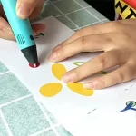 Nahaufnahme einer Hand, die mit einem blauen Marker auf einem weißen Papier gelbe und rote Kreise zeichnet, auf einem grünen Fliesenboden.