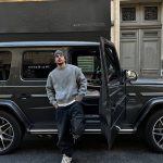 Ein junger Mann in grauem Pullover und Mütze lehnt an einem schwarzen Luxus-SUV, geöffnetes Tür, urbaner Hintergrund, stilvoll und modern.
