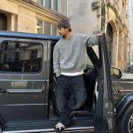 Ein junger Mann in casual Kleidung steht an einem schwarzen Auto in einer urbanen Umgebung.