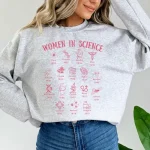 Graues Sweatshirt mit Illustrationen von Frauen in der Wissenschaft und ihren Namen, das den weiblichen Beitrag zur Forschung und wissenschaftlichen Entdeckung feiert.